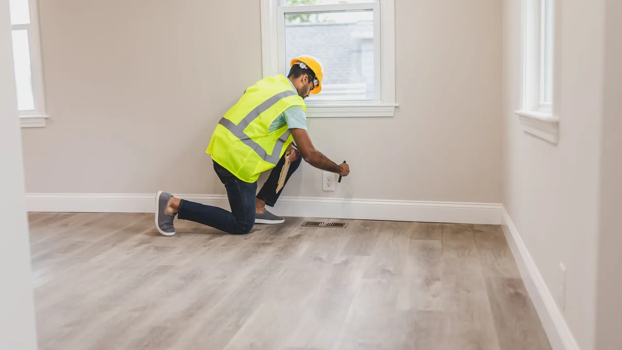 An inspector in a hard hat and high-visibility vest kneels to check a wall at floor level in a bright, empty room.